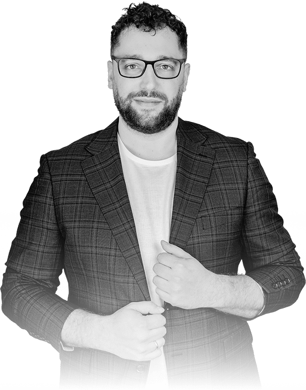 Mężczyzna z kręconymi włosami i okularami ma na sobie kraciastą marynarkę na zwykłym t-shircie, zwrócony do przodu i dopasowujący marynarkę obiema rękami. Tło jest gładkie i białe.